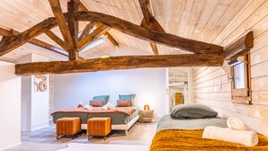 Un dormitorio con vigas de madera a la vista, una cama con manta verde, dos otomanas y un ventanal grande.