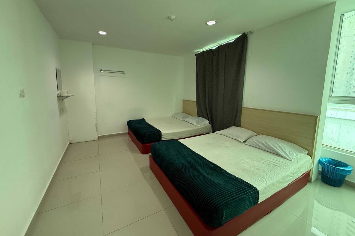 A hotel room with two beds, a small table, a mirror, a window with curtains, and a blue laundry basket.