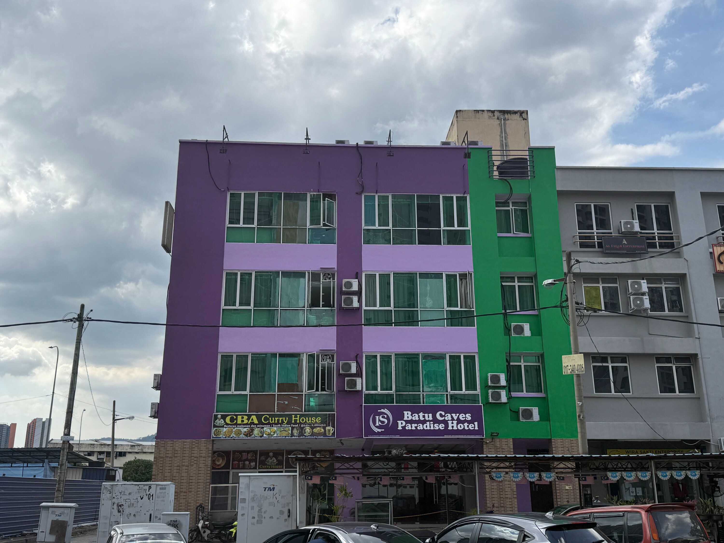 Un edificio de varios pisos con fachada morada y verde, que muestra la señalización de 'Batu Caves Paradise Hotel' y 'CBA Curry House'.