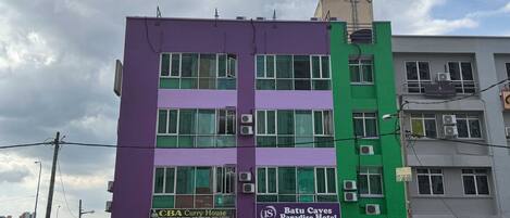 Un edificio de varios pisos con fachada morada y verde, que muestra la señalización de 'Batu Caves Paradise Hotel' y 'CBA Curry House'.