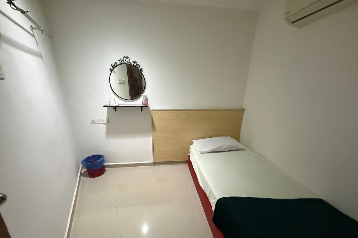 A small, clean room with a single bed, a bedside table, a mirror, and a small shelf with toiletries.