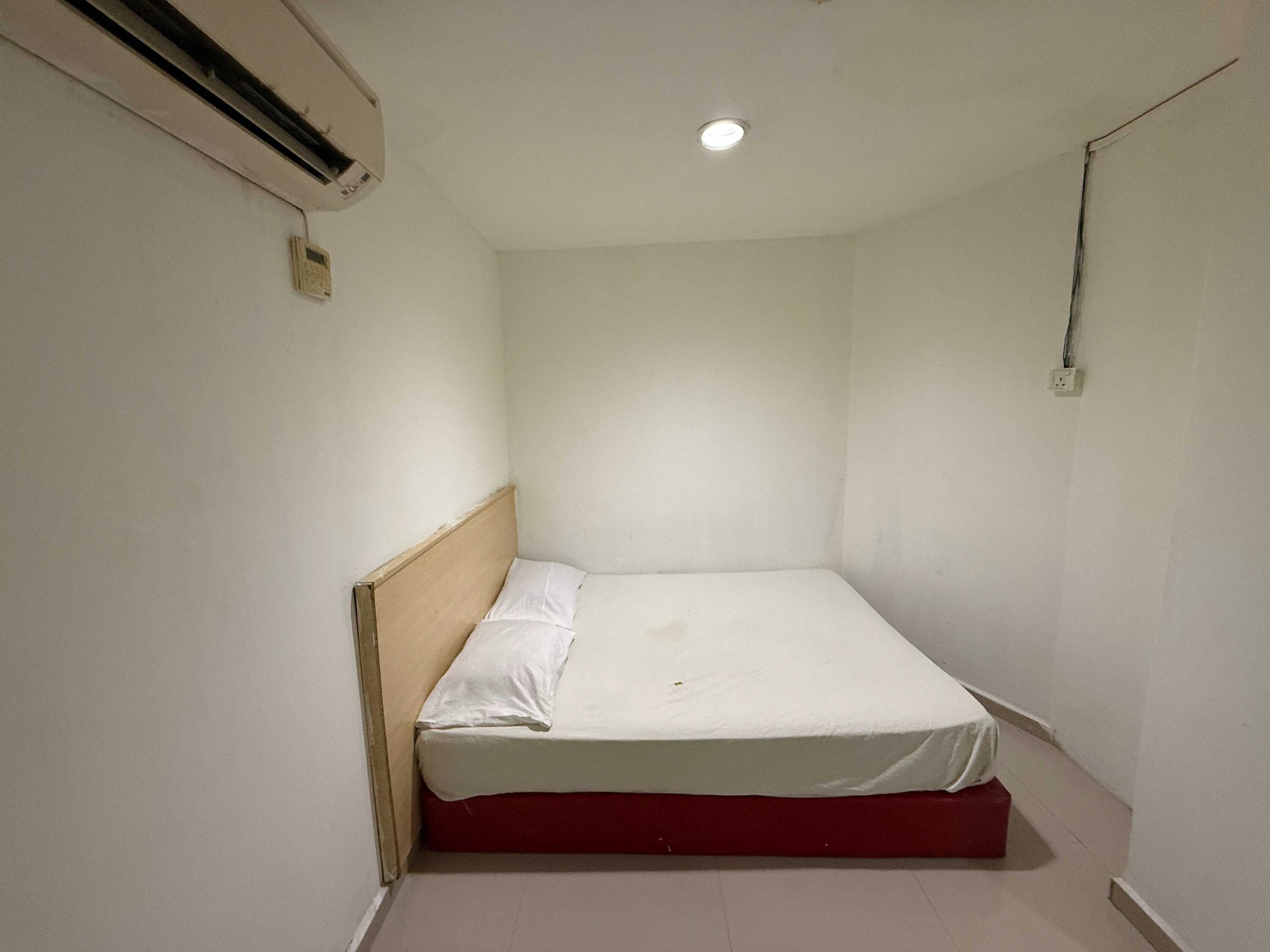 Una habitación pequeña con una cama individual, cabecera de madera, colchón blanco y base roja. Incluye una luz de techo y un equipo de aire acondicionado de pared.