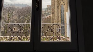 En vy genom ett fönster med en smidesjärnsbalkong, en kyrka med ett högt torn och en klocka, och en molnig himmel.