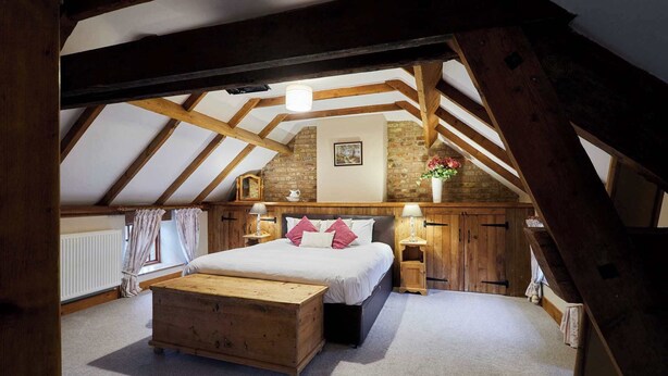 A bedroom with a sloped ceiling, wooden beams, a bed with pillows, a nightstand, a dresser, a radiator, and a brick wall with a picture. - Host & Stay | The Grainary (Bridlington)
