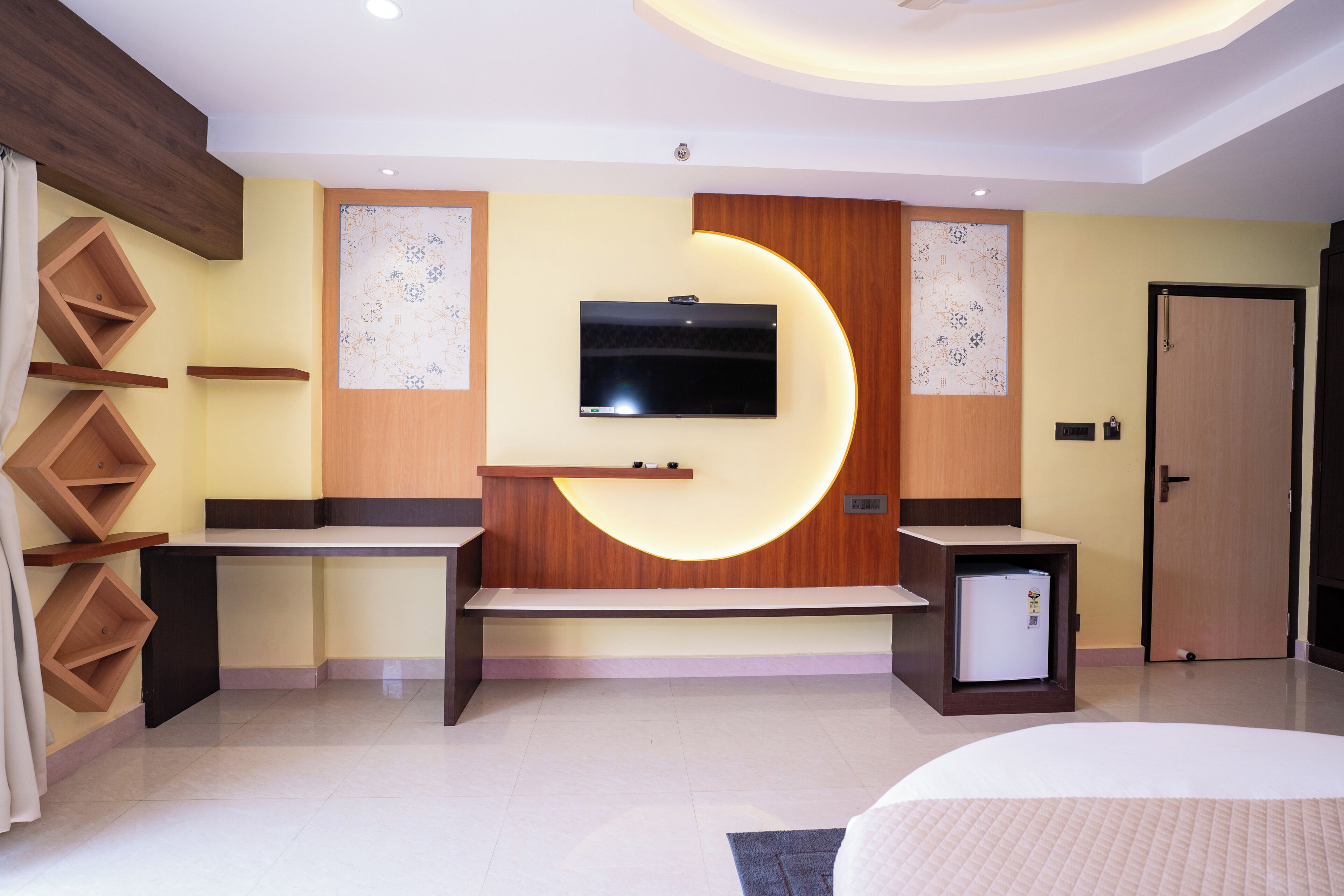 A modern hotel room with a flat-screen TV mounted on the wall, a built-in bench, wooden shelving units, and a small refrigerator.