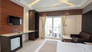 Een moderne hotelkamer met een bed, een bureau met een televisie, een kleine koelkast en een balkon met een tafel en stoelen.