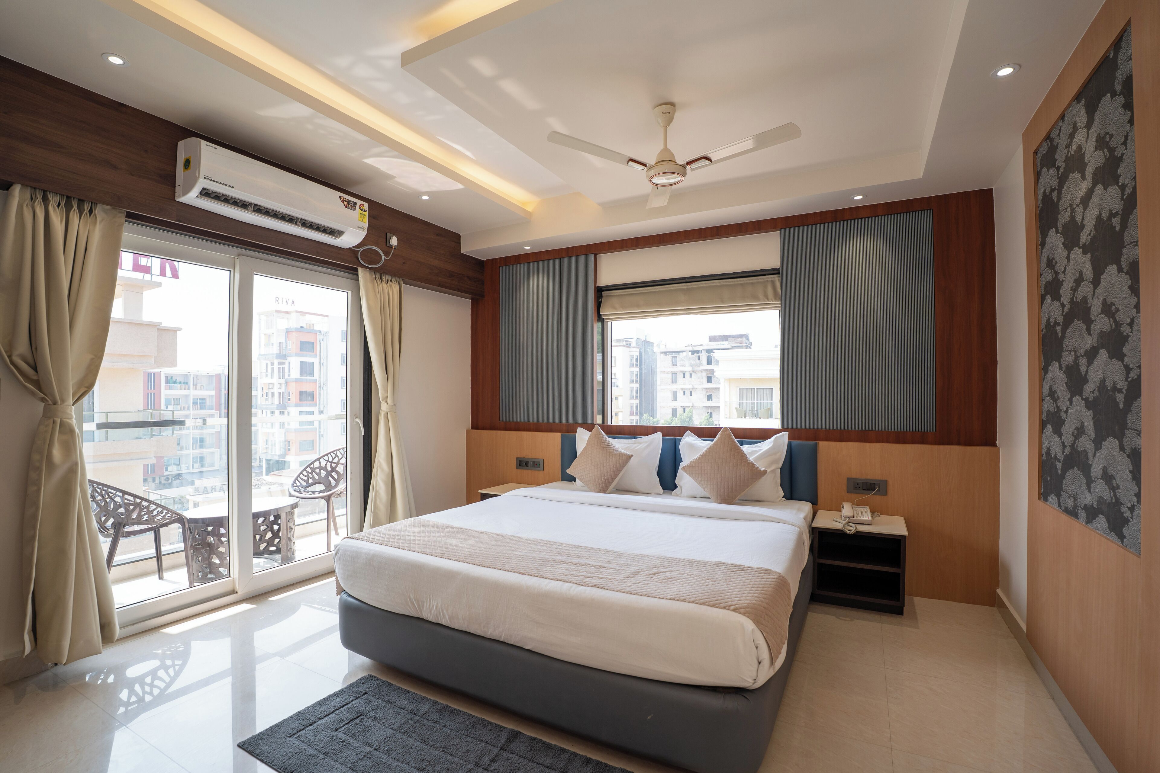 Una habitación de hotel moderna con una cama grande, un ventilador de techo, un equipo de aire acondicionado, un balcón con vistas a la ciudad y una pequeña mesa con un teléfono.