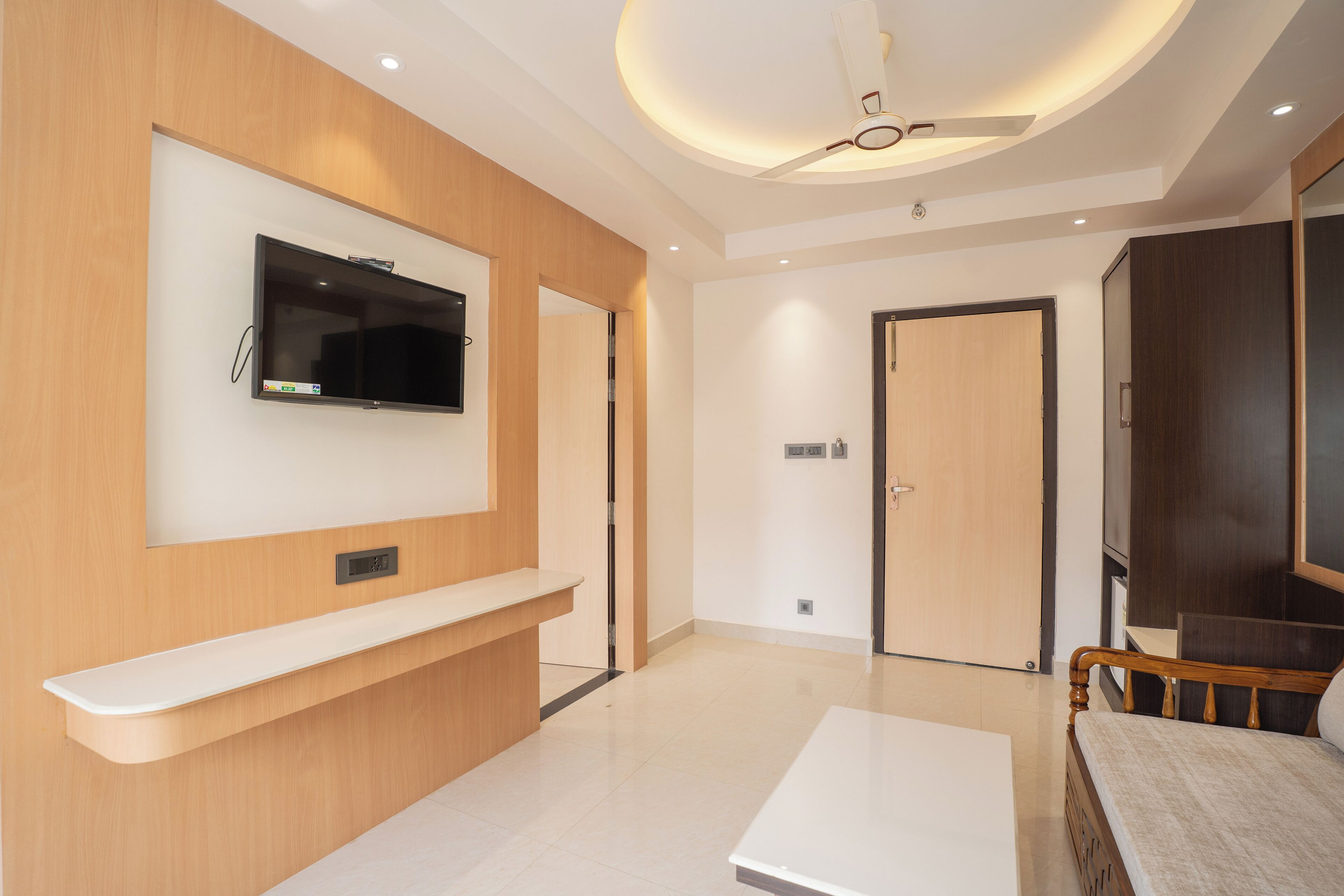 Une chambre d’hôtel moderne équipée d’un téléviseur à écran plat fixé au mur, d’un banc en bois, d’un lit avec une tête de lit en bois et d’un ventilateur de plafond.