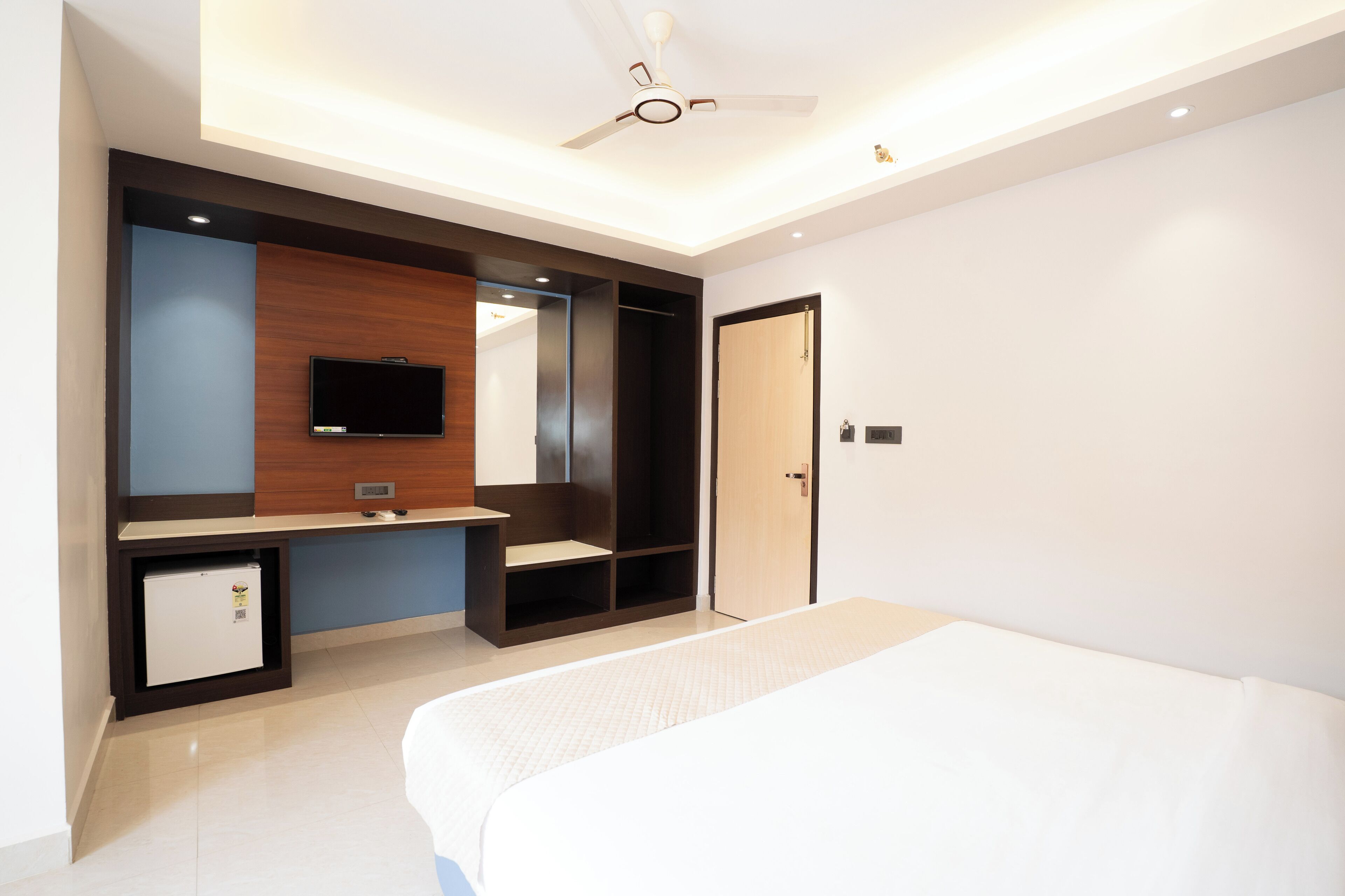 A modern hotel room with a bed, a TV, a small fridge, a wooden cabinet, and a ceiling fan.