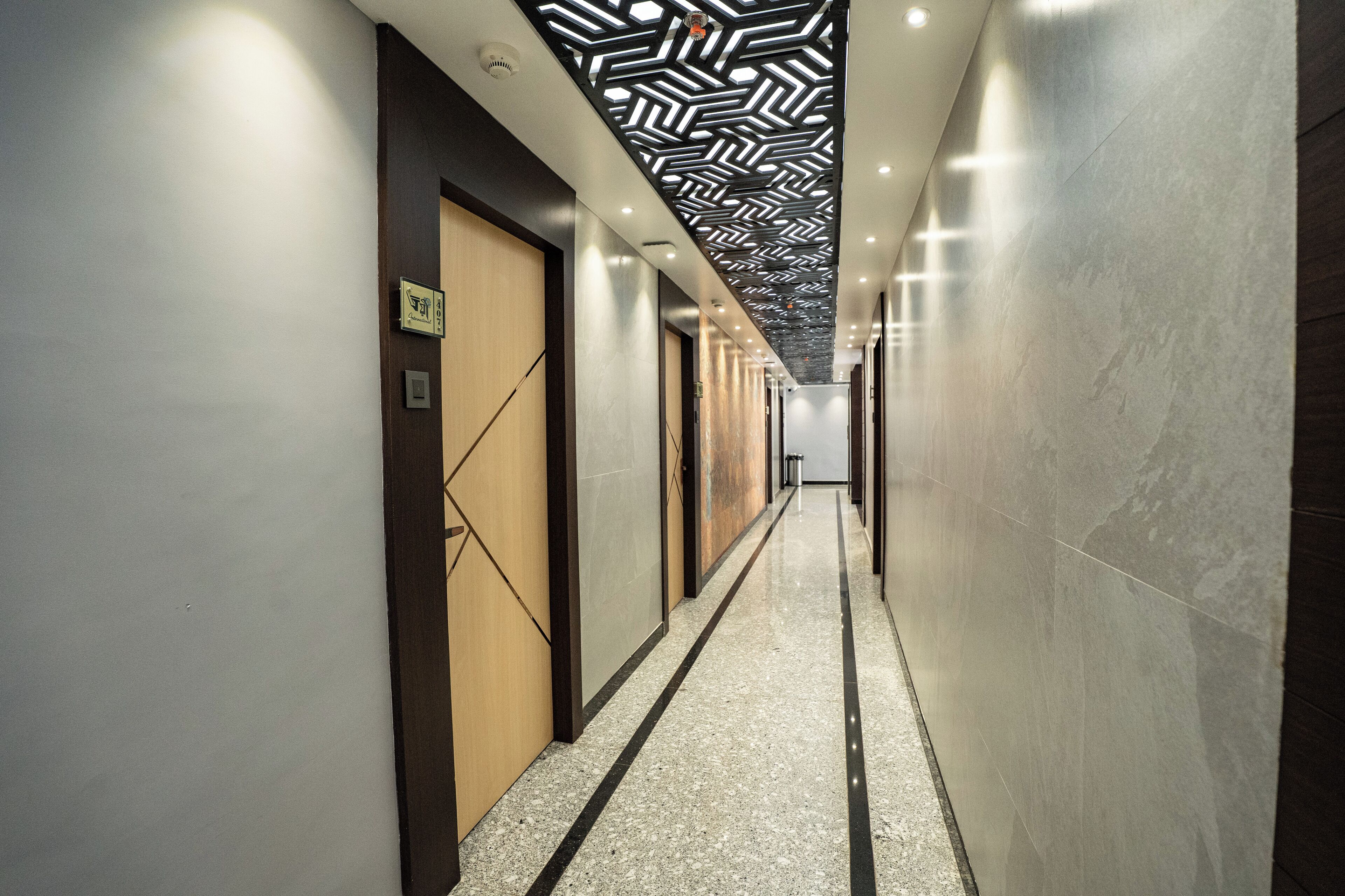A modern hotel corridor with a geometric patterned ceiling, marble flooring, and wooden doors.