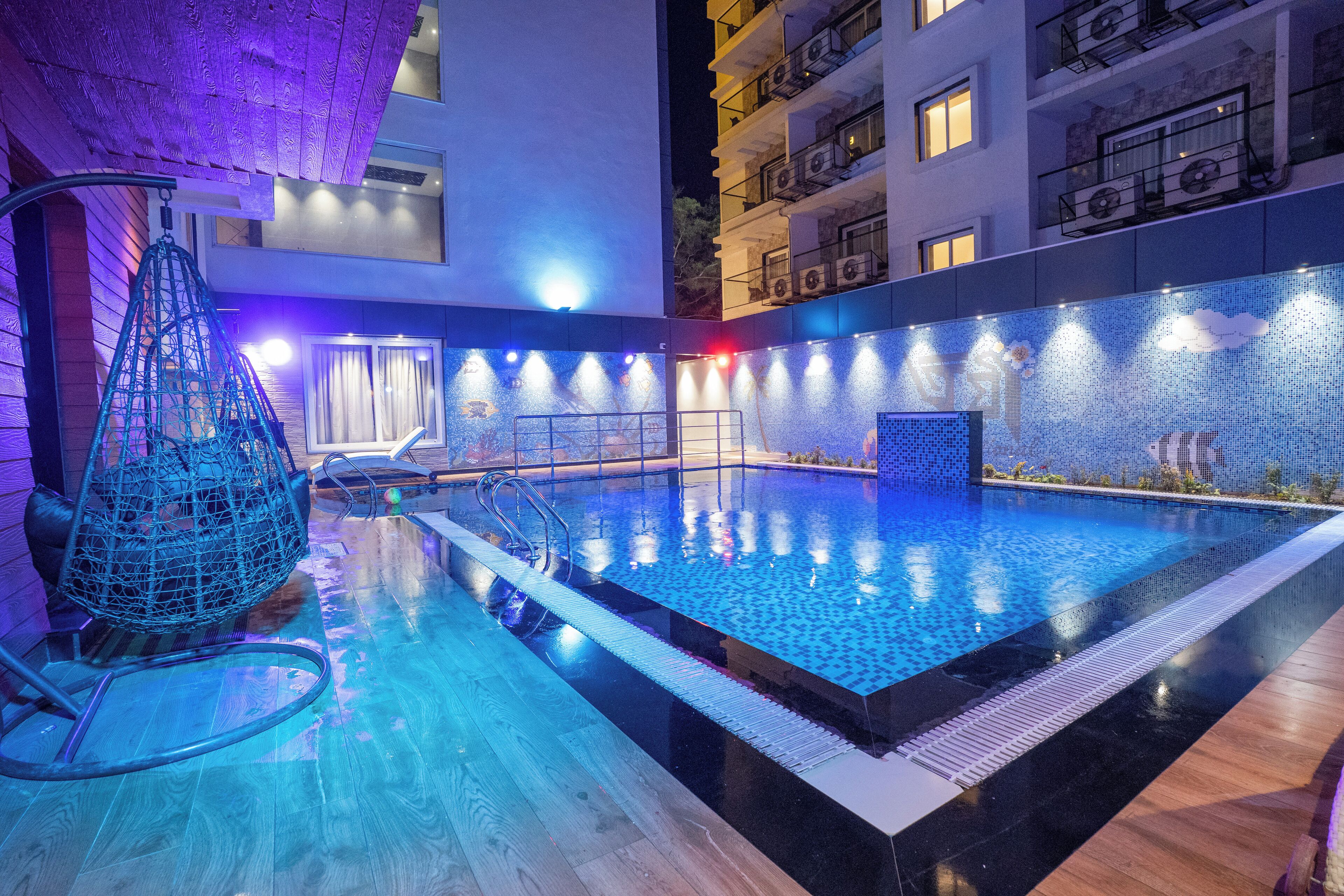 Une piscine intérieure moderne, dotée d’une eau cristalline, entourée d’une terrasse en bois et agrémentée d’un mur décoratif recouvert de mosaïques.