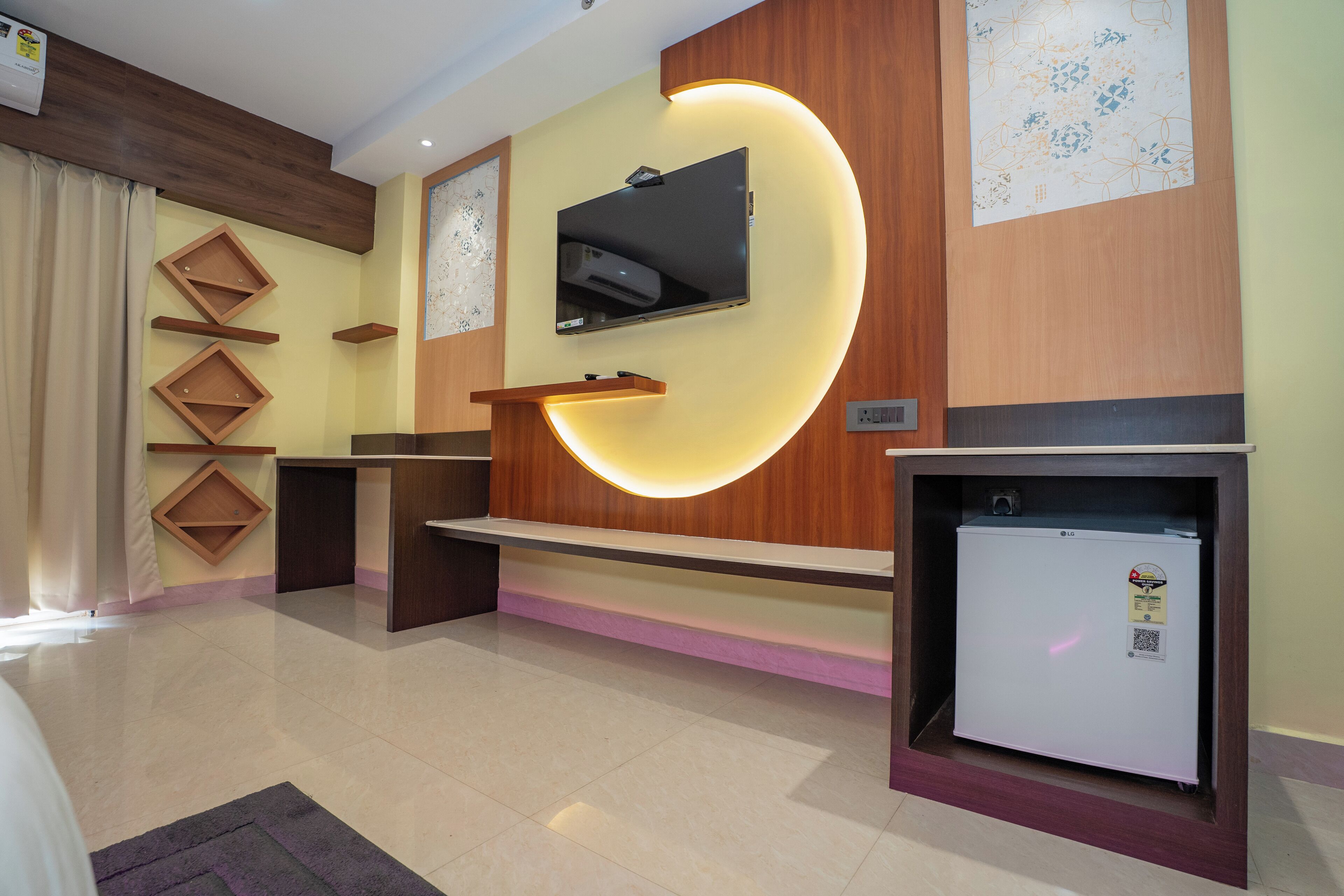 A hotel room with a flat-screen TV mounted on a wooden wall, a small refrigerator, and a wooden shelf with a built-in TV.