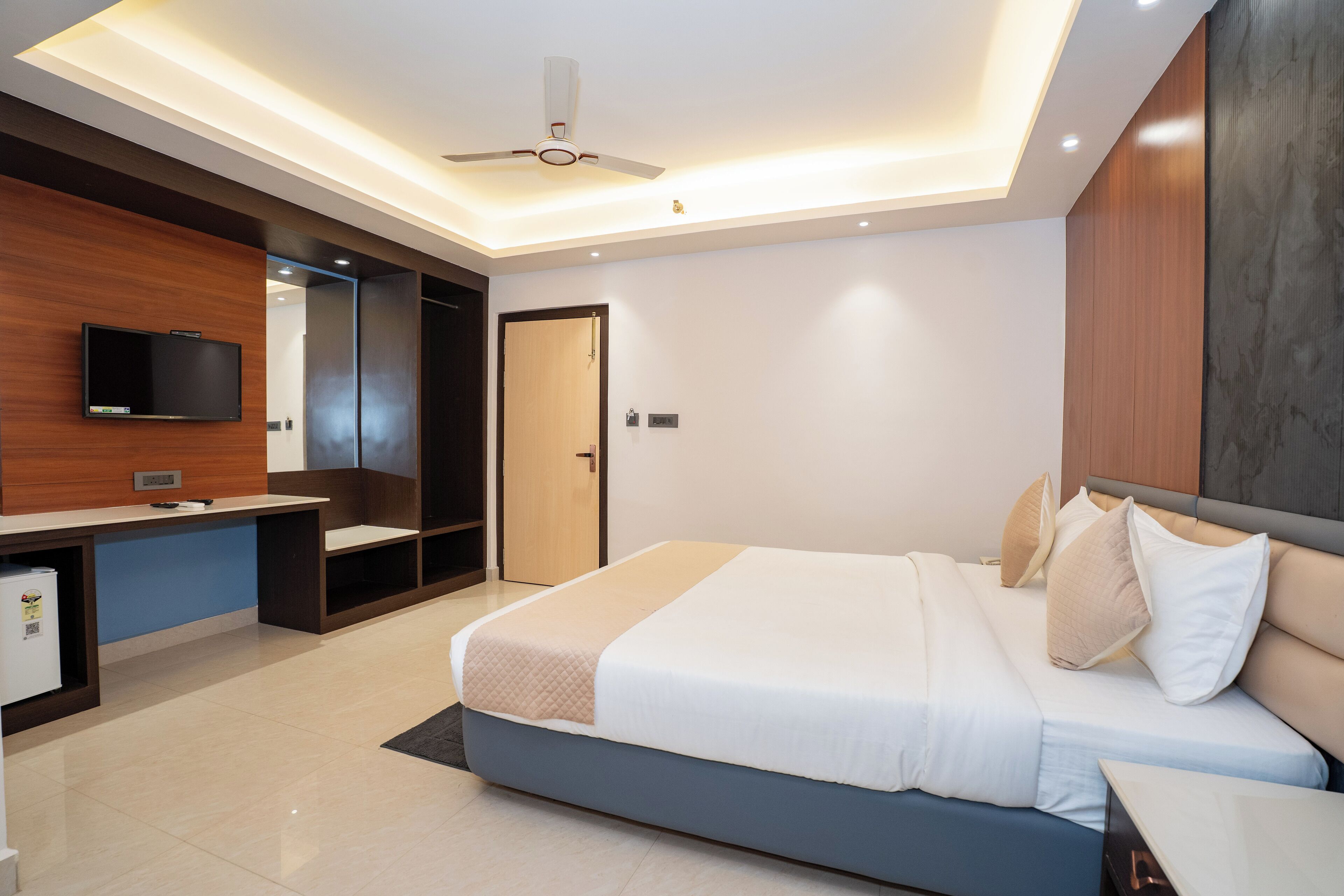 Una habitación de hotel moderna con cama, mesita de noche, televisor integrado, armario y ventilador de techo.