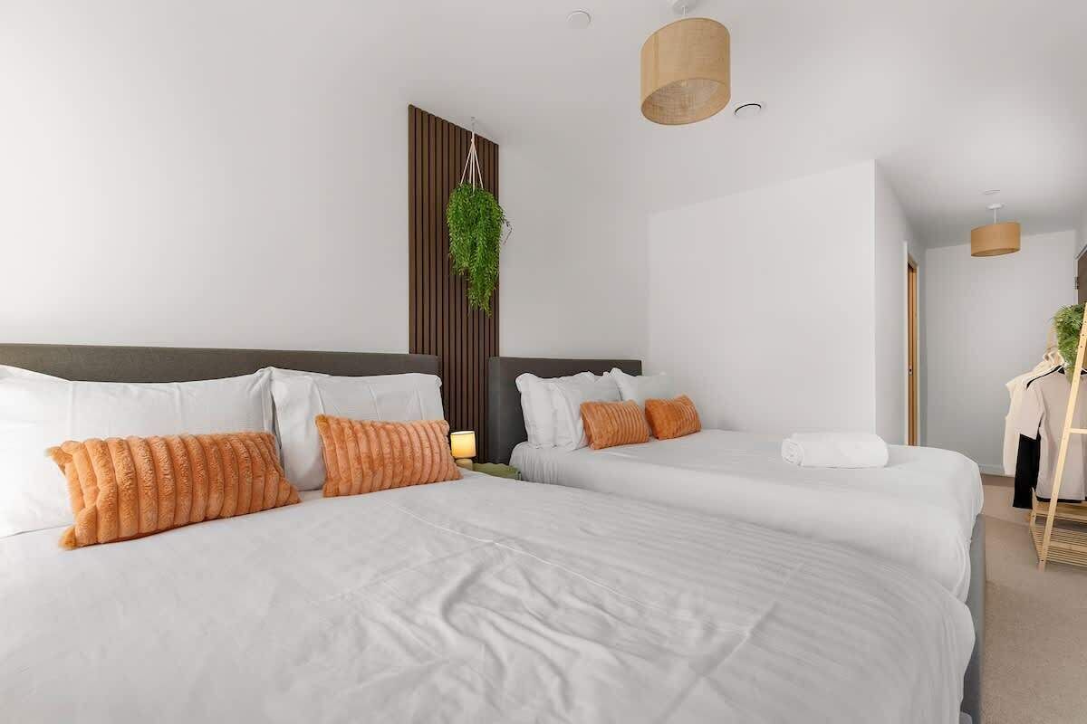 A hotel room with two beds, white bedding, orange pillows, a hanging plant, and a wooden headboard.