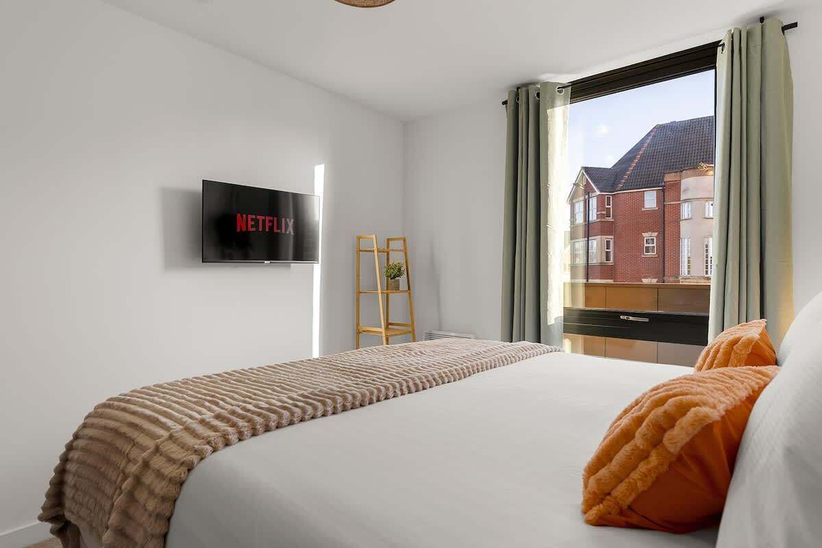 A hotel room with a bed, a TV mounted on the wall displaying 'NETFLIX', a window with curtains, and a view of buildings outside.