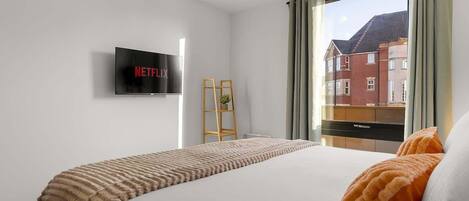 A hotel room with a bed, a TV mounted on the wall displaying 'NETFLIX', a window with curtains, and a view of buildings outside.