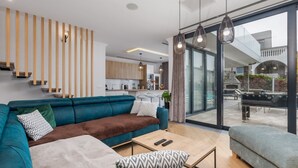 Egy modern nappali, melyben egy kék kanapé, egy dohányasztal és egy szürke karosszék található. A háttérben egy konyha látható, ahonnan egy lépcső vezet a felső szintre.