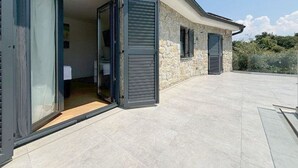 Egy modern ház, kőburkolatos homlokzattal, nagy üvegajtóval, amely egy belső helyiségbe vezet, és egy kiépített teraszrésszel.