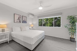Een slaapkamer met een groot bed, twee nachtkastjes met lampen, een plafondventilator, een raam met uitzicht op bomen, en een potplant.
