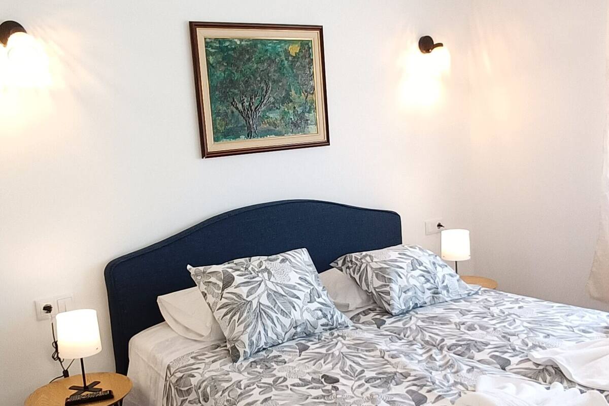 Un lit bien fait, recouvert d’une couvre-lit à motifs floraux, une table de chevet avec une lampe, un climatiseur fixé au mur et un tableau encadré au-dessus du lit.