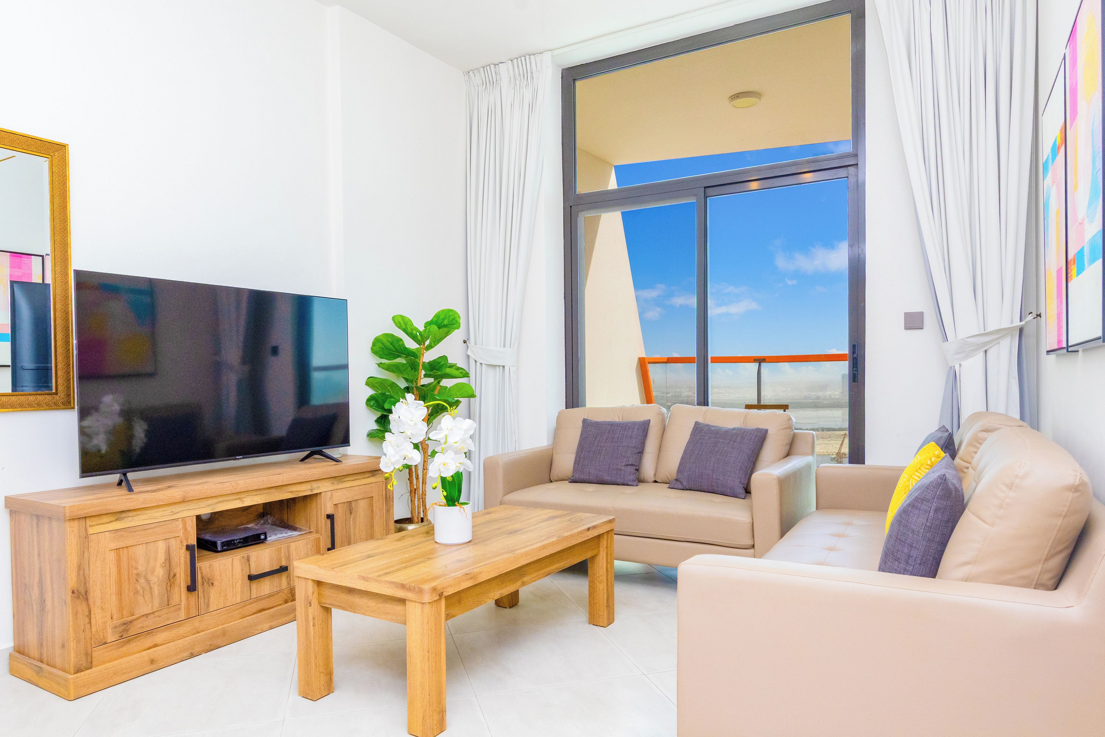 A modern living room with a flat-screen TV, wooden furniture, a coffee table, and a large window with curtains.