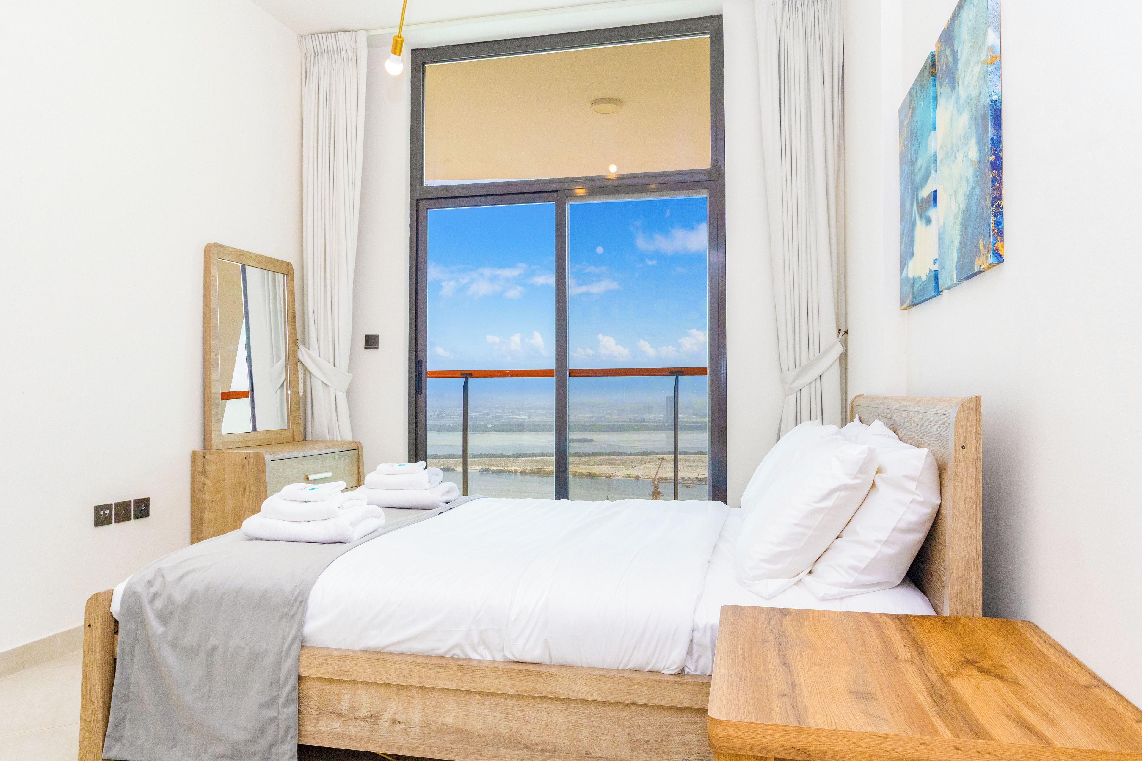 A hotel room with a large window, a bed with white linens, a wooden headboard, a bedside table, a mirror, and a painting on the wall.