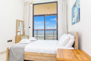 A hotel room with a large window, a bed with white linens, a wooden headboard, a bedside table, a mirror, and a painting on the wall. - Bloomfields 1BR - Binghatti Avenue - Jaddaf (Dubai)