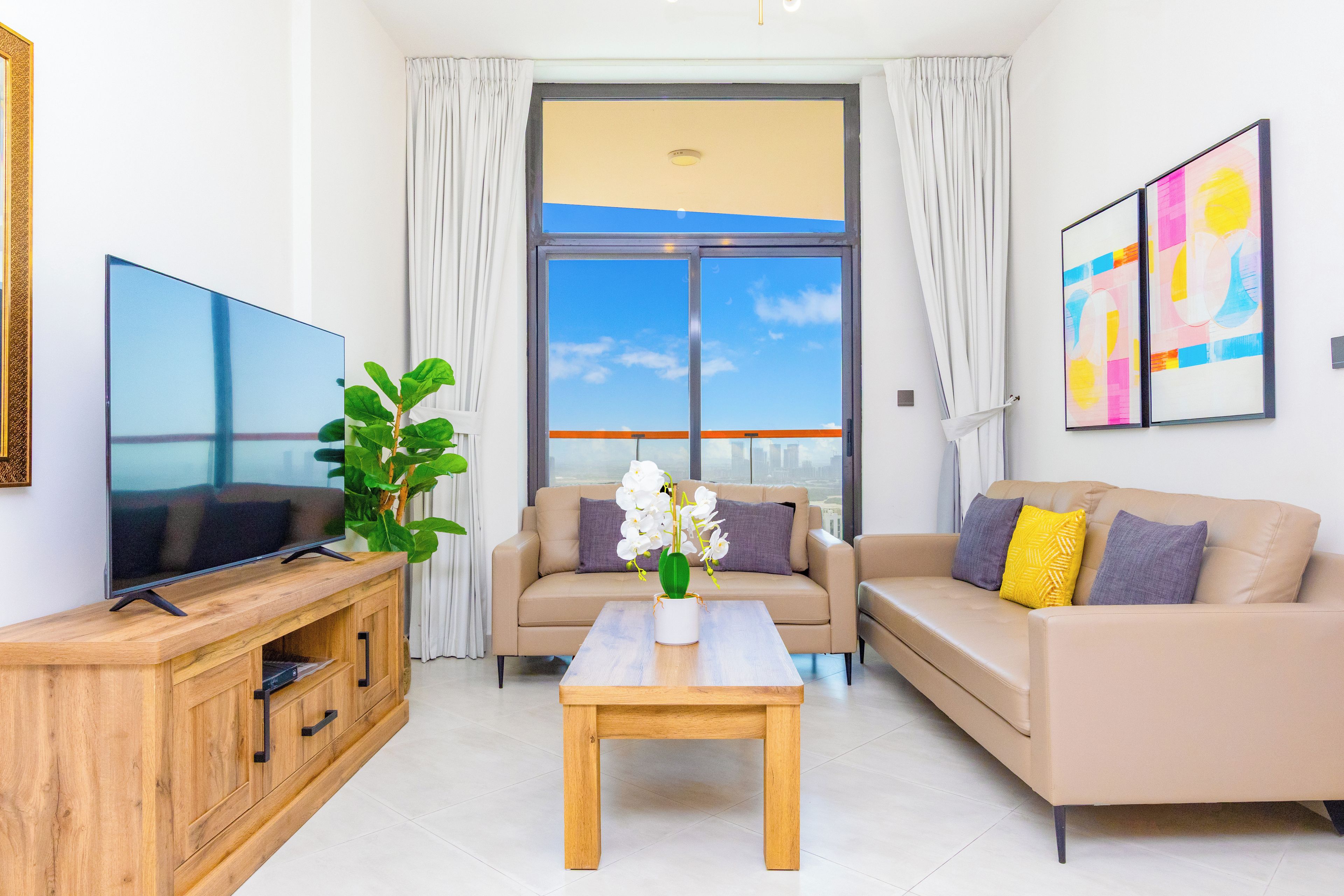 Ein modernes Wohnzimmer mit einem Flachbildfernseher, einer Holzkommode, einem beigefarbenen Sofa mit Kissen, einem Couchtisch und einem großen Fenster mit Vorhängen.