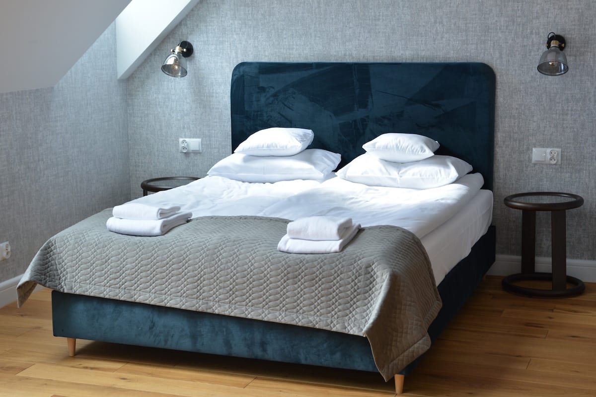 Una cama bien tendida con sábanas blancas y colcha gris, dos mesitas de noche y un techo inclinado con tragaluz.
