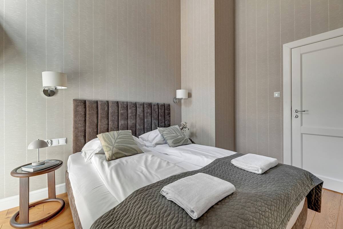 Una cama bien hecha con sábanas blancas, una colcha gris acolchada y dos toallas dobladas. Junto a la cama, una mesita de madera redonda con una lámpara.