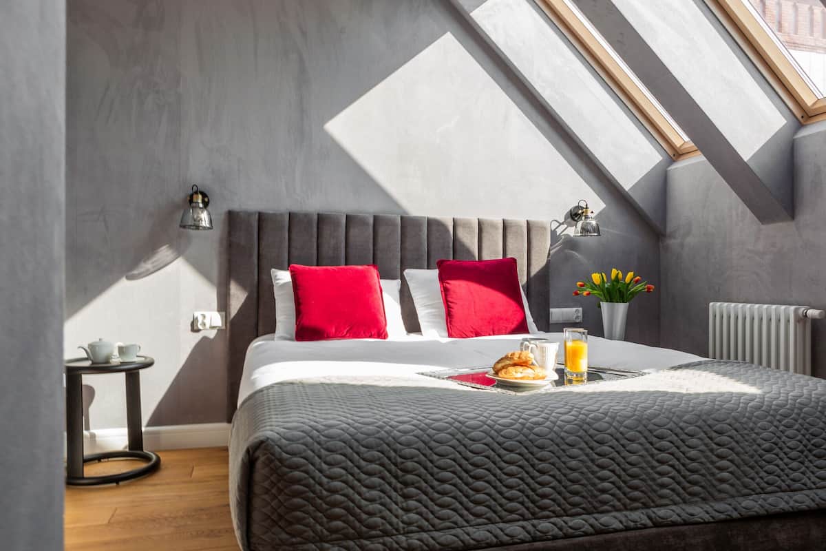 Un dormitorio moderno con colcha acolchada gris, almohadas rojas y blancas, una mesita de noche con lámpara, una pequeña mesa con tetera y una bandeja de desayuno.