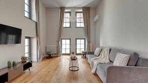 Een ruime woonkamer met een grijze bank, een ronde salontafel en een flatscreen televisie aan de muur.