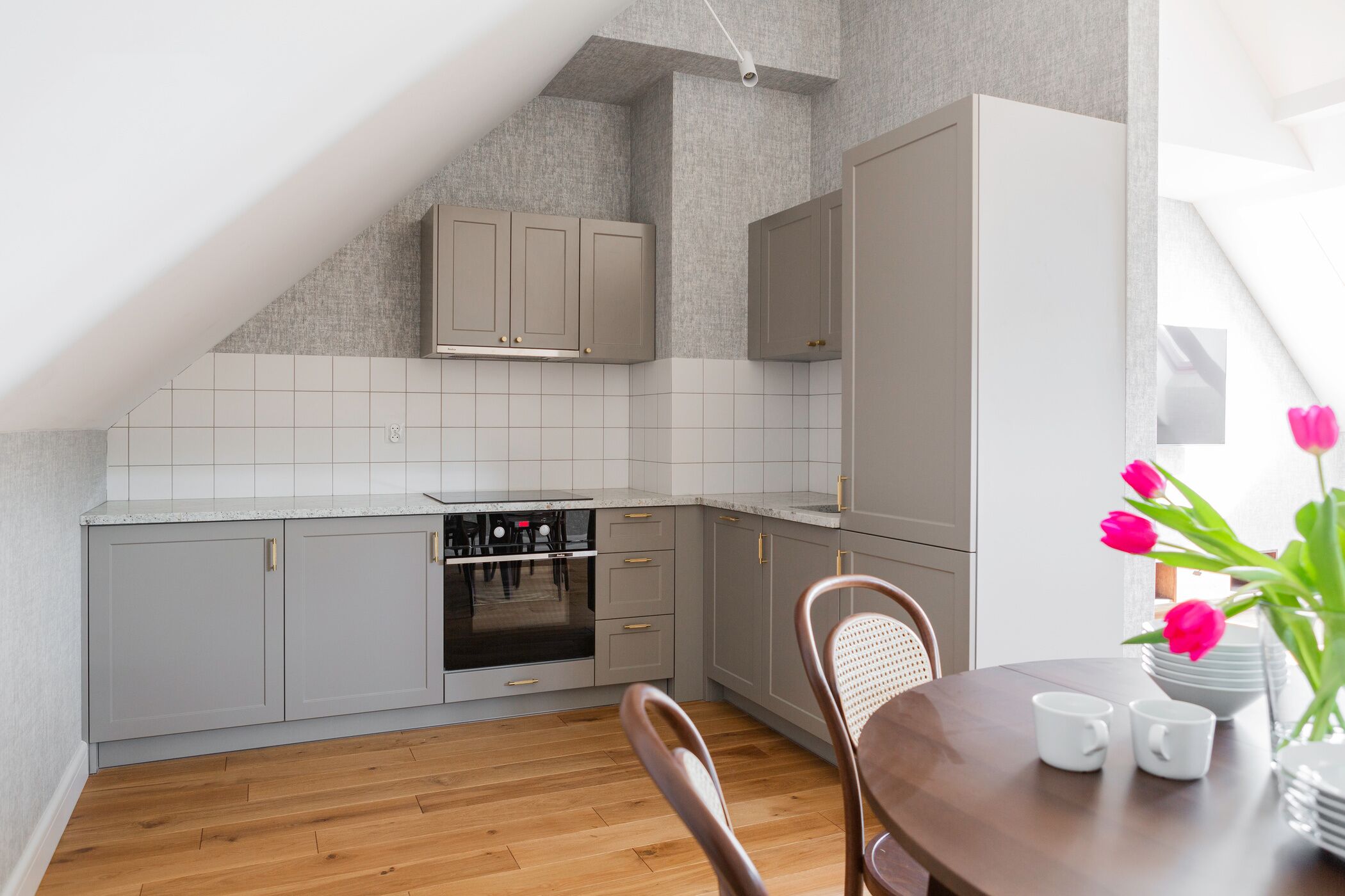 Een moderne keuken met grijze kastjes, een inbouwoven en een eethoek met een houten tafel en stoelen.
