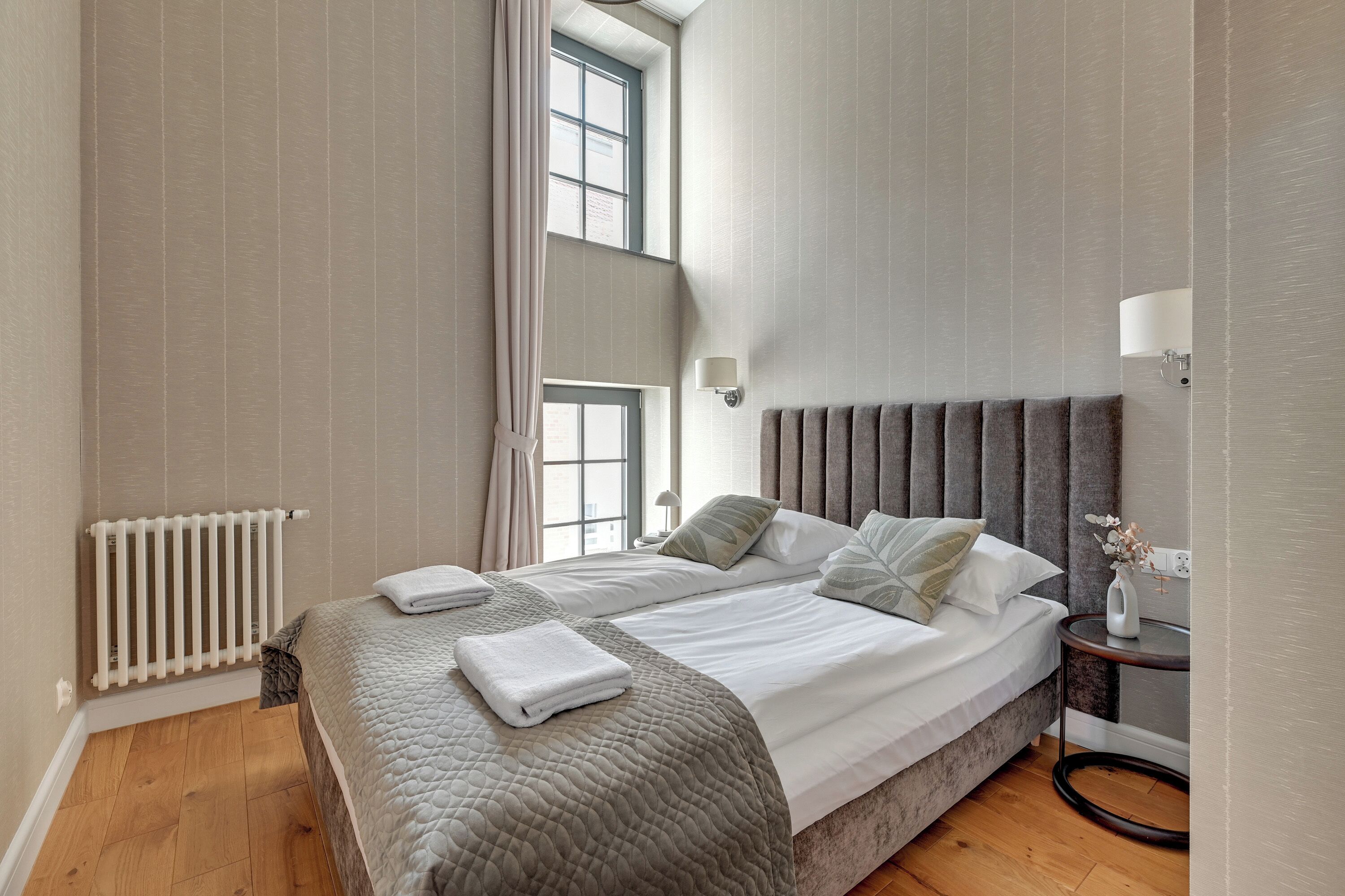 A double bed with a grey headboard, a bedside table with a lamp, and a window with curtains.