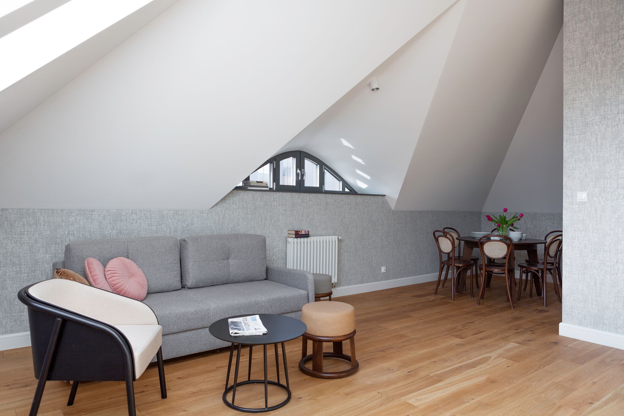 Un loft moderne avec un plafond en pente, un canapé gris, une chaise noire, une petite table ronde et un coin repas avec des chaises et une table en bois.