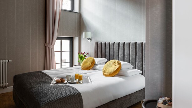 Een net opgemaakt bed met een dienblad vol ontbijtartikelen, een bedlampje en een raam met gordijnen.