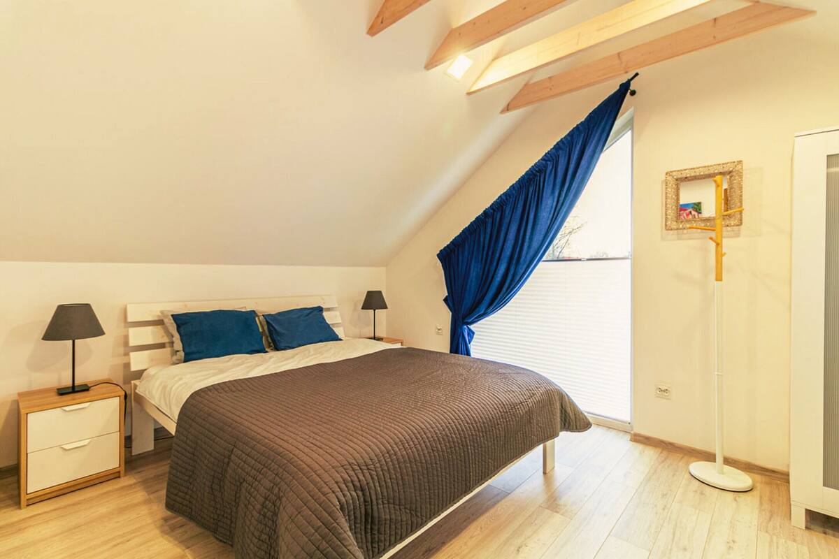 Quarto com cama, mesas de cabeceira, abajur, janela com cortinas e um quadro na parede.
