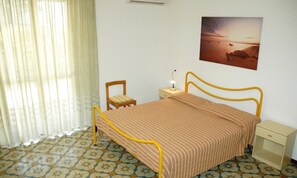 A bedroom with a bed, a chair, a nightstand, a lamp, a picture on the wall, and a window with curtains. - Practical Studio in Lignano Pineta (Lignano Sabbiadoro)