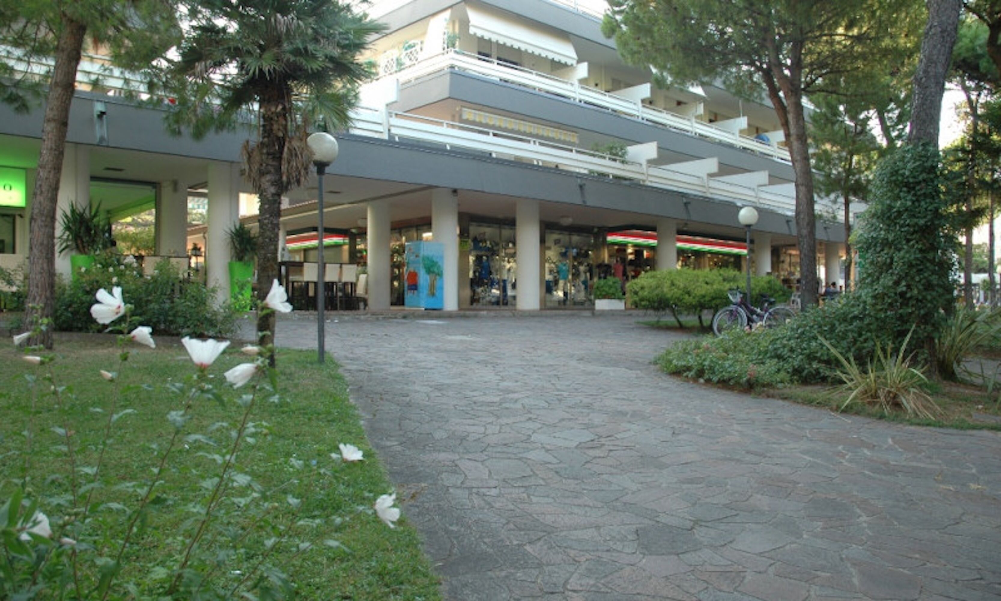 Un centro comercial de varios niveles con un paseo adoquinado de piedra, palmeras y una bicicleta estacionada cerca de la entrada.