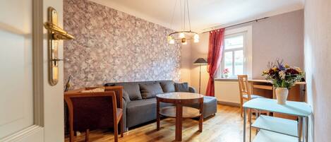 A living room with a sofa, a round wooden table, a dining table with chairs, a radiator, and a window with curtains.