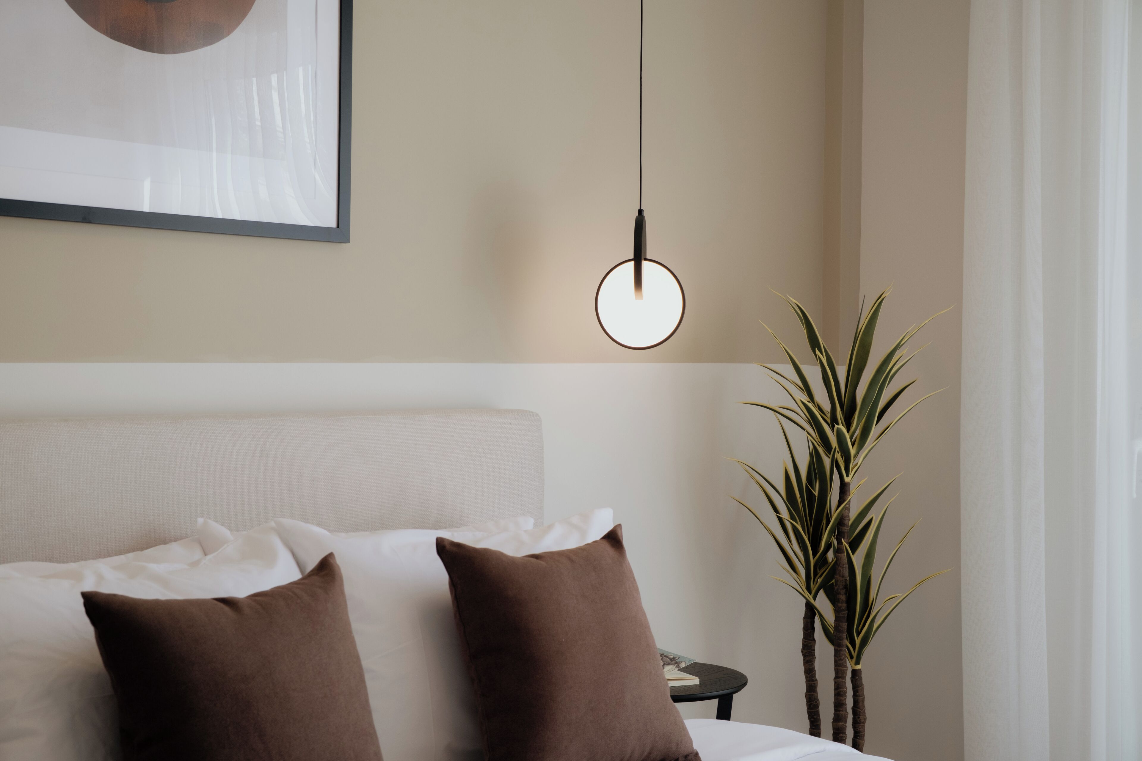 A neatly made bed with white and brown pillows, a hanging light fixture, and a potted plant beside the bed.