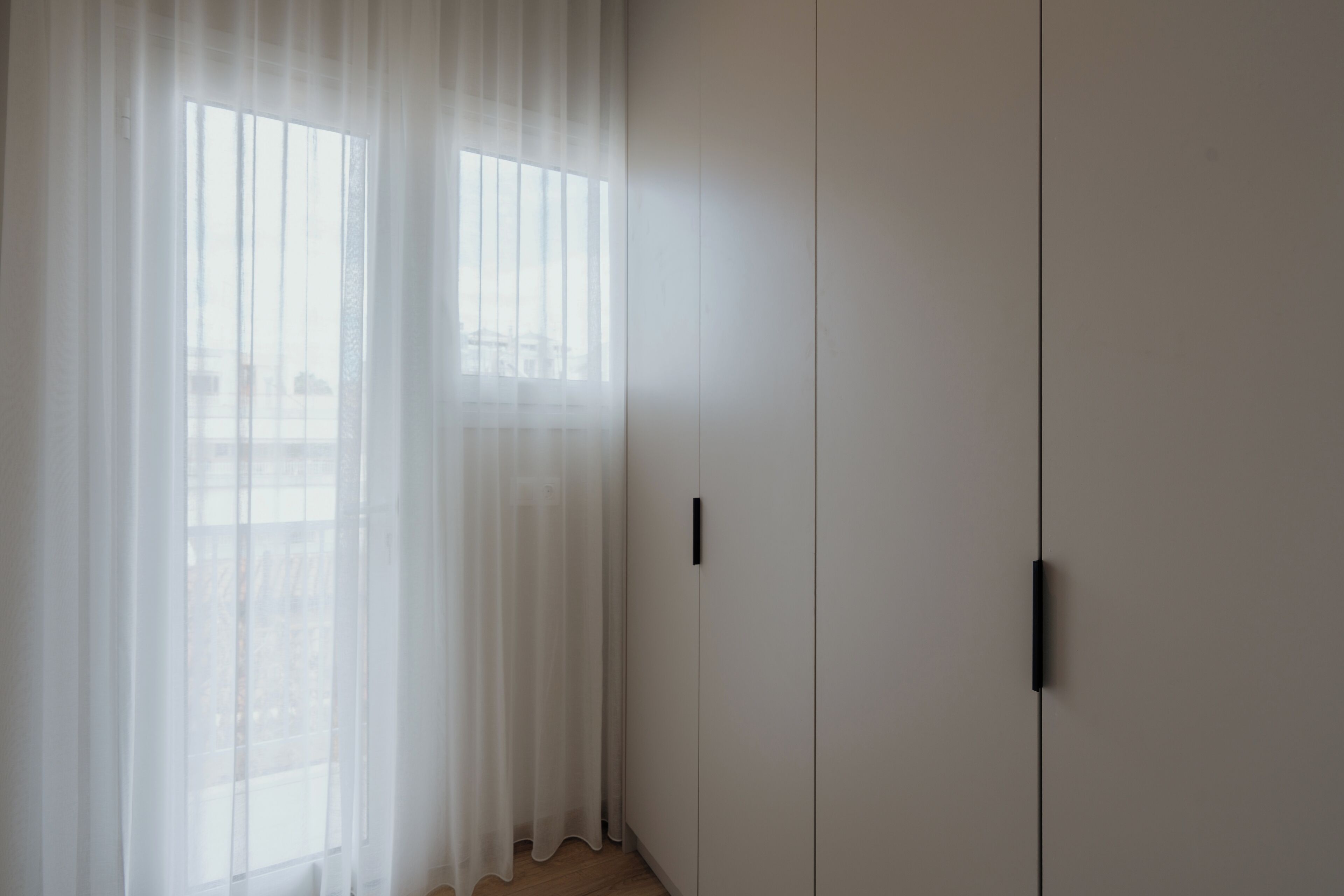 A room with a large window covered by sheer curtains, a white wardrobe, and a visible door handle.