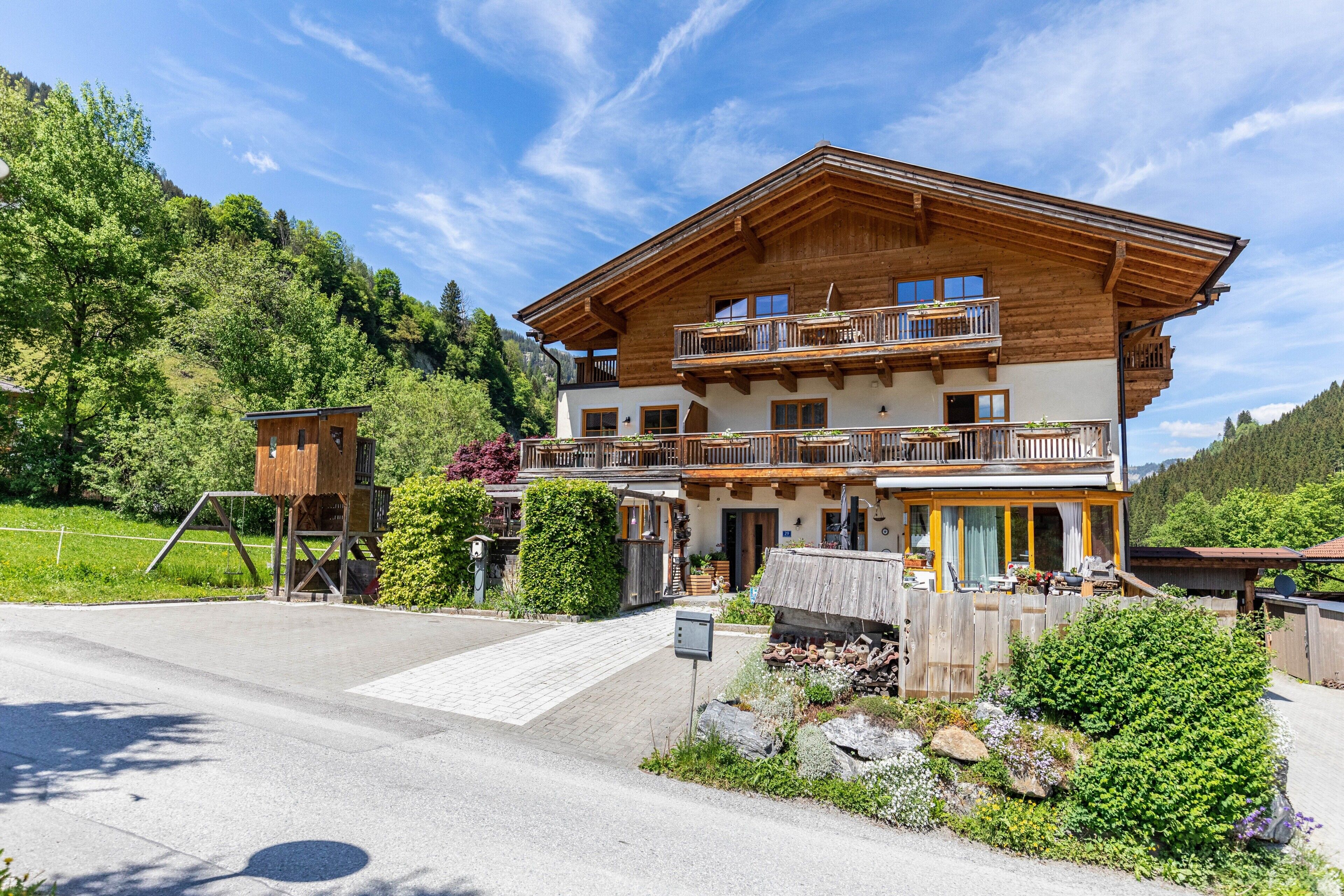 A two-story wooden chalet with balconies, a wooden deck, and a mountain backdrop.