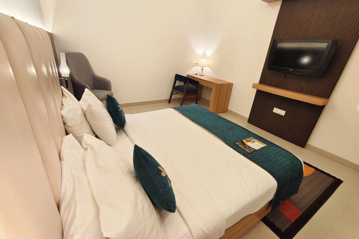 Een netjes opgemaakt bed met witte en teal beddengoed, een bureau met een lamp en een aan de muur bevestigde televisie.