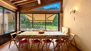 A dining area with a table set for six, wooden chairs with red cushions, a large window with a view of greenery, and a brick wall. - Cascine di Villa Schella with Pool – Relaxing Oasis for Groups and Families (Ovada)
