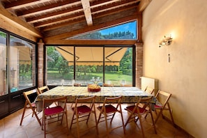 A dining area with a table set for six, wooden chairs with red cushions, a large window with a view of greenery, and a brick wall.