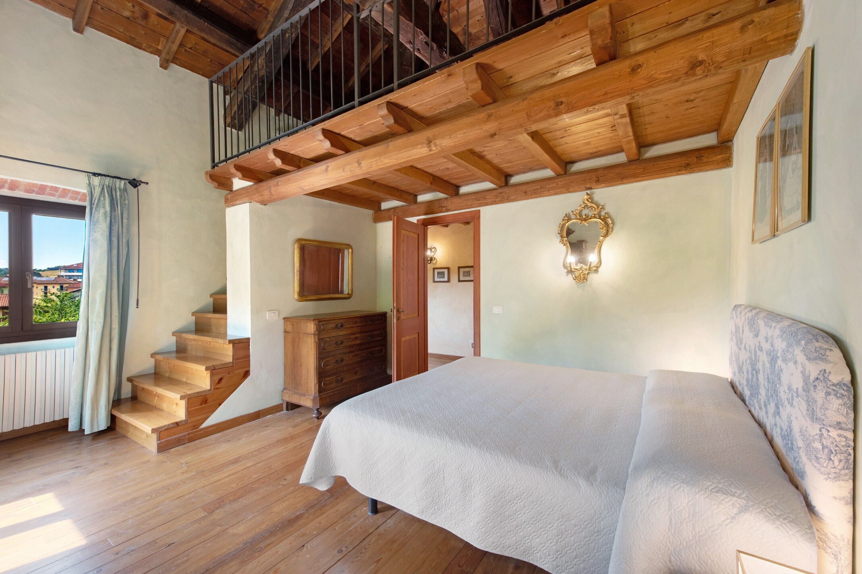 A bedroom with a wooden staircase, a bed, a dresser, and a window with curtains.