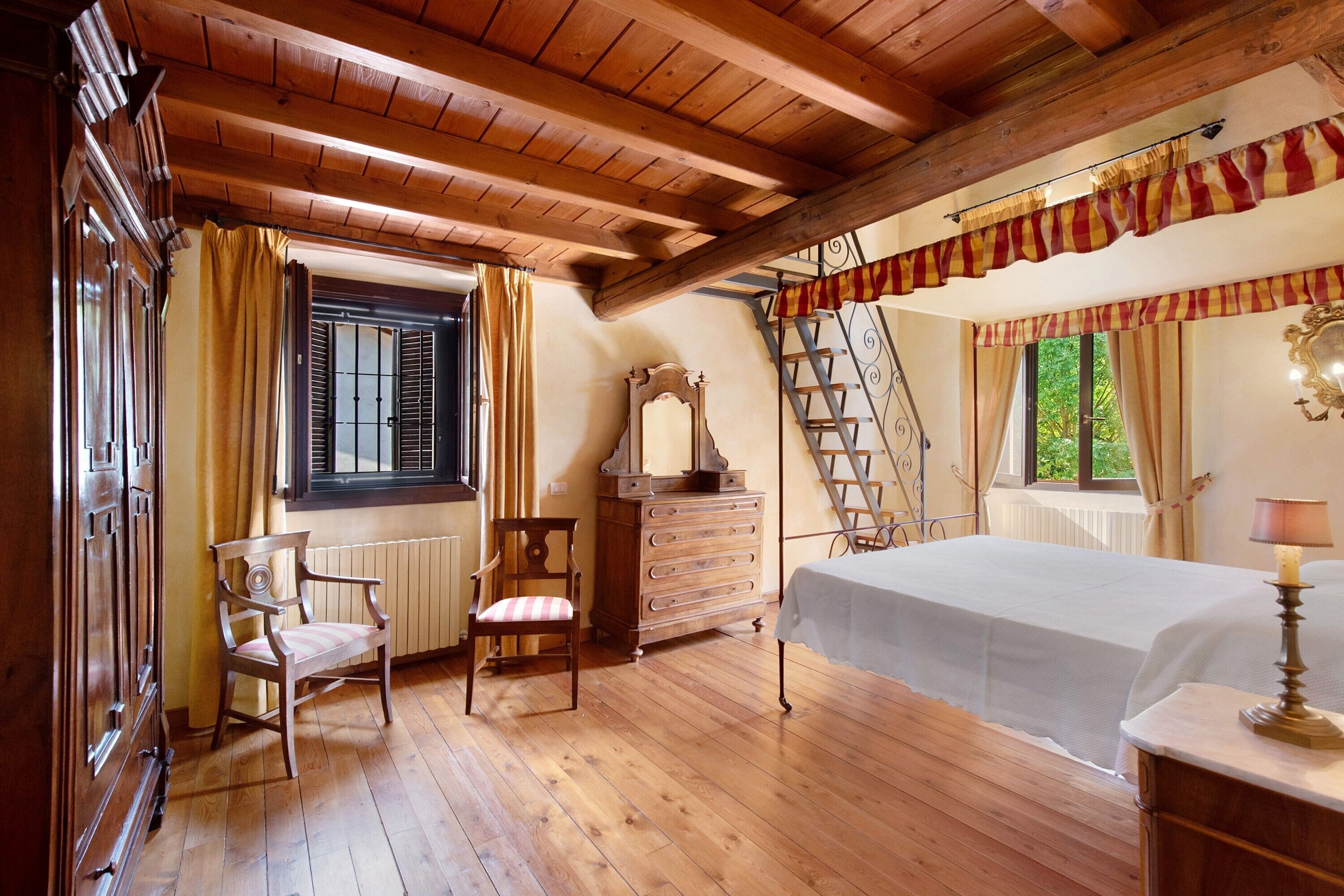 Ein Zimmer mit Holzböden, einem Bett, einem Schminktisch, einem Stuhl, einem Fenster mit Rollläden und einer steilen Treppe.