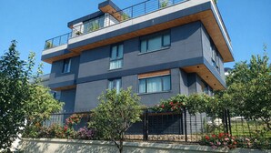 A modern multi-story building with balconies, large windows, and a garden area with flowers. - AHSA VILLA WONDERFOUL SEA VIEW AND SWIMMING POOL (İstanbul)