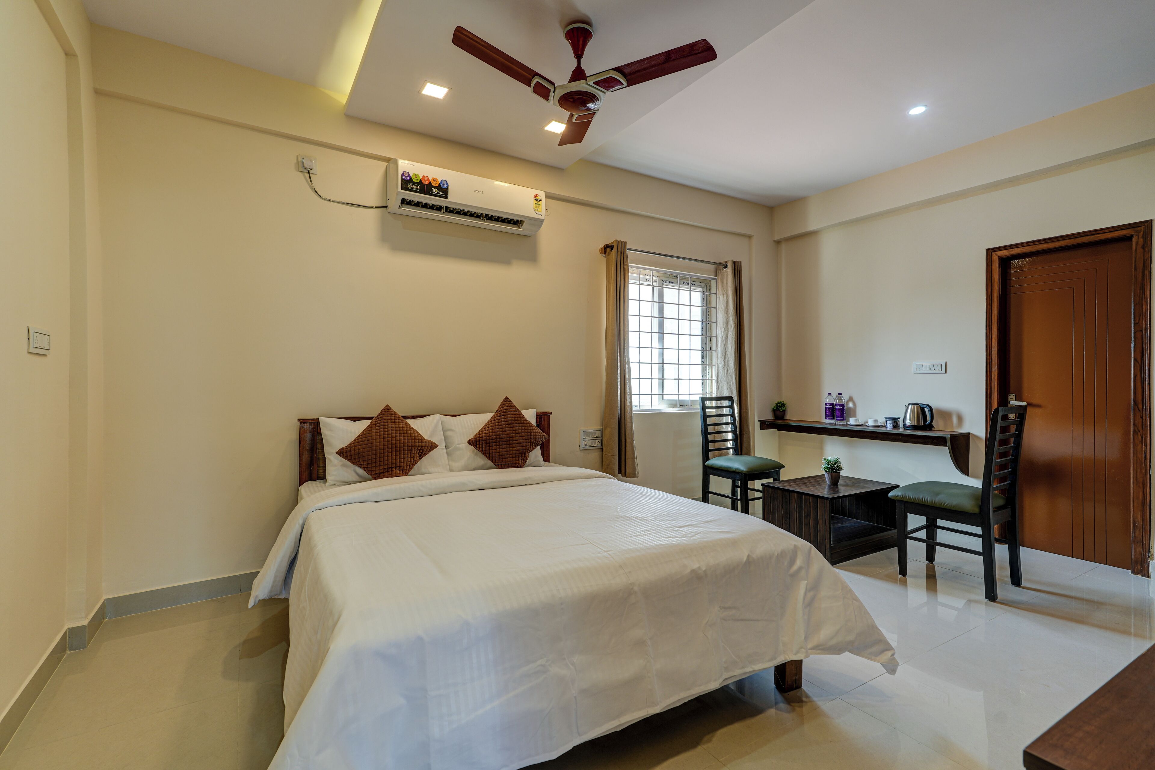 Habitación de hotel con cama, escritorio, silla, ventilador de techo, aire acondicionado y ventana con cortinas.